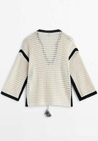 Cardigan léger en tricot ajouré crème avec des accents noirs. Présente des manches trois-quarts et un détail à frange à l'arrière.