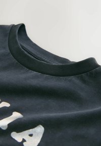 Gros plan d'un t-shirt noir à col rond côtelé avec un texte blanc imprimé partiellement visible sur le tissu.