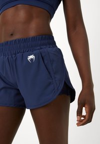 Close-up van een persoon die marineblauwe sportbroekjes draagt met een wit logo en een bijpassende sportbeha, staand tegen een effen achtergrond.