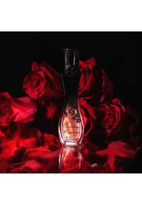 Flacone di profumo ricurvo con etichetta "Christina Aguilera By Night" tra vivaci rose rosse e petali su sfondo scuro.