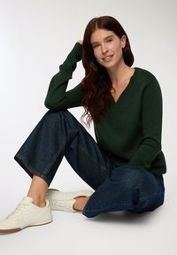 Zielony sweter dzianinowy z dekoltem w kształcie litery V, w połączeniu z szerokimi, ciemnoniebieskimi dżinsami i białymi trampkami, charakteryzuje się luźnym krojem i ściągaczami przy mankietach.