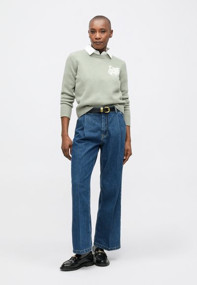 Suéter verde claro con un cuello blanco, que cuenta con un bolsillo texturizado. Combinado con unos vaqueros de mezclilla azul de cintura alta y zapatos negros.