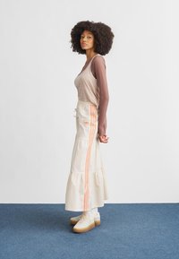 Šviesiai ruda marškinėliai ilgomis rankovėmis, derinami su kreminės spalvos sluoksniuota sijonu su oranžinėmis juostelėmis. Modelis dėvi baltus batus su smėlio spalvos pado detalėmis. Lygūs audinių paviršiai.