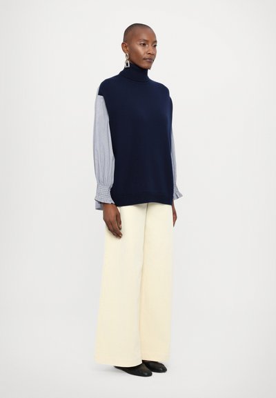 Navy knit sleeveless turtleneck top with light blue striped long sleeves, paired with light cream wide-leg trousers and black shoes.