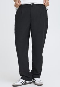 Pantalon noir à coupe ample, avec plis sur le devant et fermeture par bouton. Texture lisse, associé à des baskets blanches et grises.