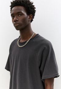 Tricou gri cu mânecă scurtă, realizat dintr-un material moale, care are o croială lejeră și margini brutale la manșete, completat de un colier din lanț de argint stratificat.