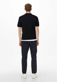 Zwarte polo met korte mouwen en geribbelde kraag, gecombineerd met donkere marineblauwe broek en witte sneakers, gezien vanuit de achterkant.