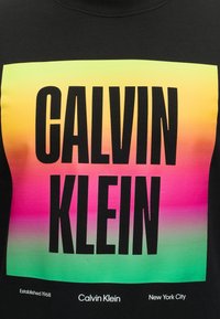 Μαύρο πουκάμισο με μεγάλο κείμενο CALVIN KLEIN σε φόντο με κλίση από κίτρινο σε ροζ και πράσινο, με μικρότερο κείμενο "Established 1968" και "New York City."