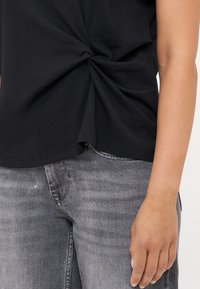 Zwarte top met korte mouwen met een geknoopte detail in de taille, gemaakt van zachte stof, gecombineerd met lichtgrijze spijkerbroek.