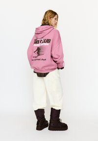 Felpa con cappuccio rosa con stampa "SKI CLUB Winter Series", vestibilità oversize, abbinata a pantaloni cargo bianchi e stivali imbottiti marroni per l'inverno.