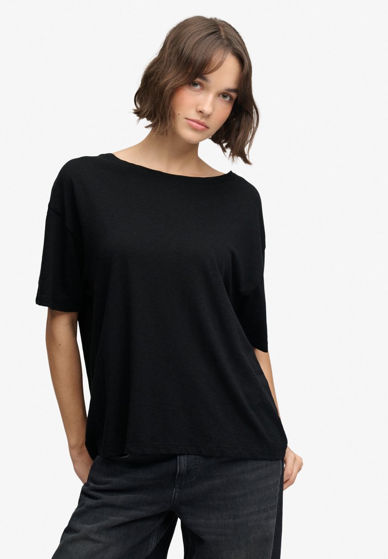 Frau mit kurzen braunen Haaren trägt ein lockeres schwarzes T-Shirt und dunkle Jeans, steht mit einer Hand in der Tasche vor einem schlichten Hintergrund.