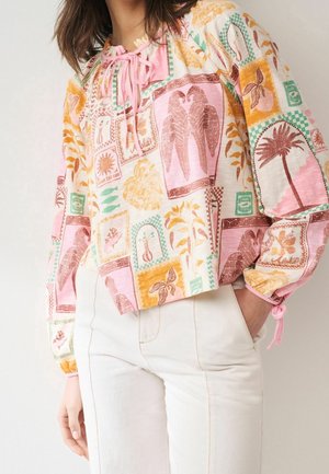 Kobieta ubrana w pastelową bluzkę typu patchwork z motywami ptaków, roślin oraz palmy, w połączeniu z białymi, wysokim stanem spodniami, z ręką w kieszeni.