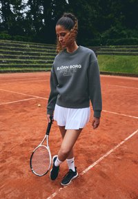 Sudadera gris con el texto "BJÖRN BORG ATHLETIC CLUB", falda de tenis blanca con pliegues, zapatos negros, sosteniendo una raqueta de tenis blanca en la cancha de arcilla.