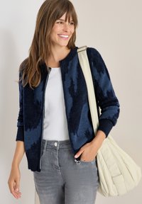 Marineblaues, flauschiges Reißverschluss-Cardigan mit abstraktem Muster, getragen über einem weißen Hemd, kombiniert mit grauen Jeans. Accessorized mit einer cremefarbenen Schultertasche.