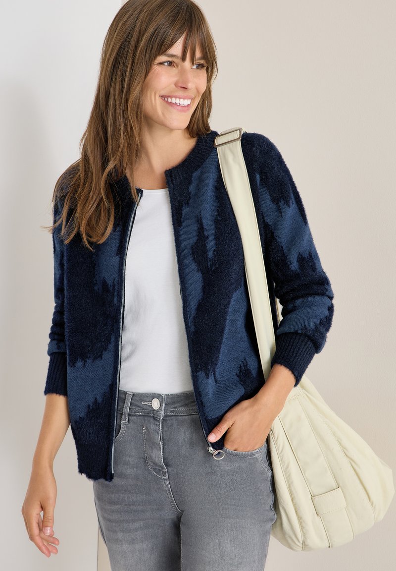 Marineblaues, flauschiges Reißverschluss-Cardigan mit abstraktem Muster, getragen über einem weißen Hemd, kombiniert mit grauen Jeans. Accessorized mit einer cremefarbenen Schultertasche.
