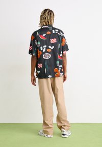 Camisa negra de manga corta con motivos coloridos de comida y bebida, combinada con pantalones beige claros y zapatillas blancas sobre un suelo verde.