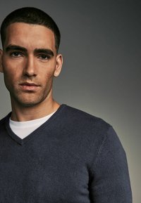 Pulover v-neck bleu marin, confecționat dintr-un material tricotat moale, purtat peste un tricou alb, având o textură netedă și un design ajustat.