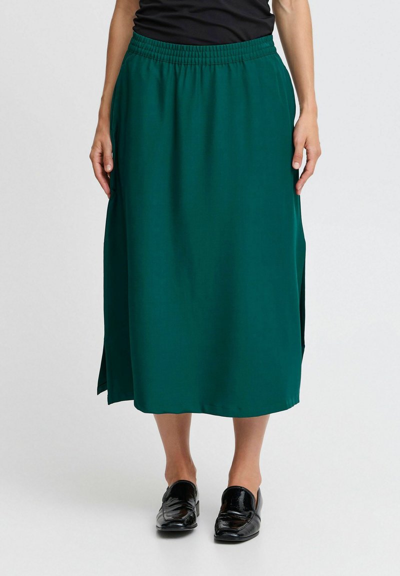Jupe midi verte en tissu lisse avec une taille élastique et des fentes latérales, associée à des chaussures noires à enfiler. Design minimaliste.