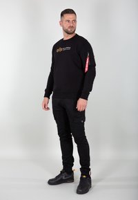 Sweatshirt noir avec logo doré, design ajusté, et poche zippée rouge ; associé avec un pantalon cargo noir et des baskets noires.