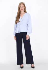 Blusa avvolgente azzurro chiaro con maniche lunghe, legata in vita, abbinata a pantaloni ampi blu navy e tacchi alti neri. Texture morbida.
