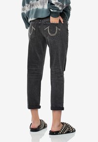 Jeans de mezclilla negra con un ajuste relajado, dobladillos acuffados y bolsillos traseros distintivos con acentos bordados. Se llevan con sandalias negras y beige.
