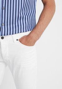 Camisa de botones a rayas en azul y marino, combinada con jeans de mezclilla blancos. La camisa cuenta con un cuello clásico y dos botones visibles en el frente.