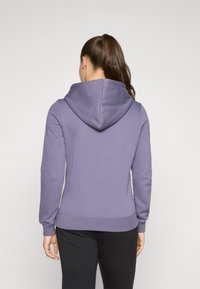 Ljubičasti hoodie s velikom kapuljačom, dugim rukavima i rebrastim manžetama. Tkanina izgleda mekano s glatkom teksturom, nošen preko tamnih hlača.