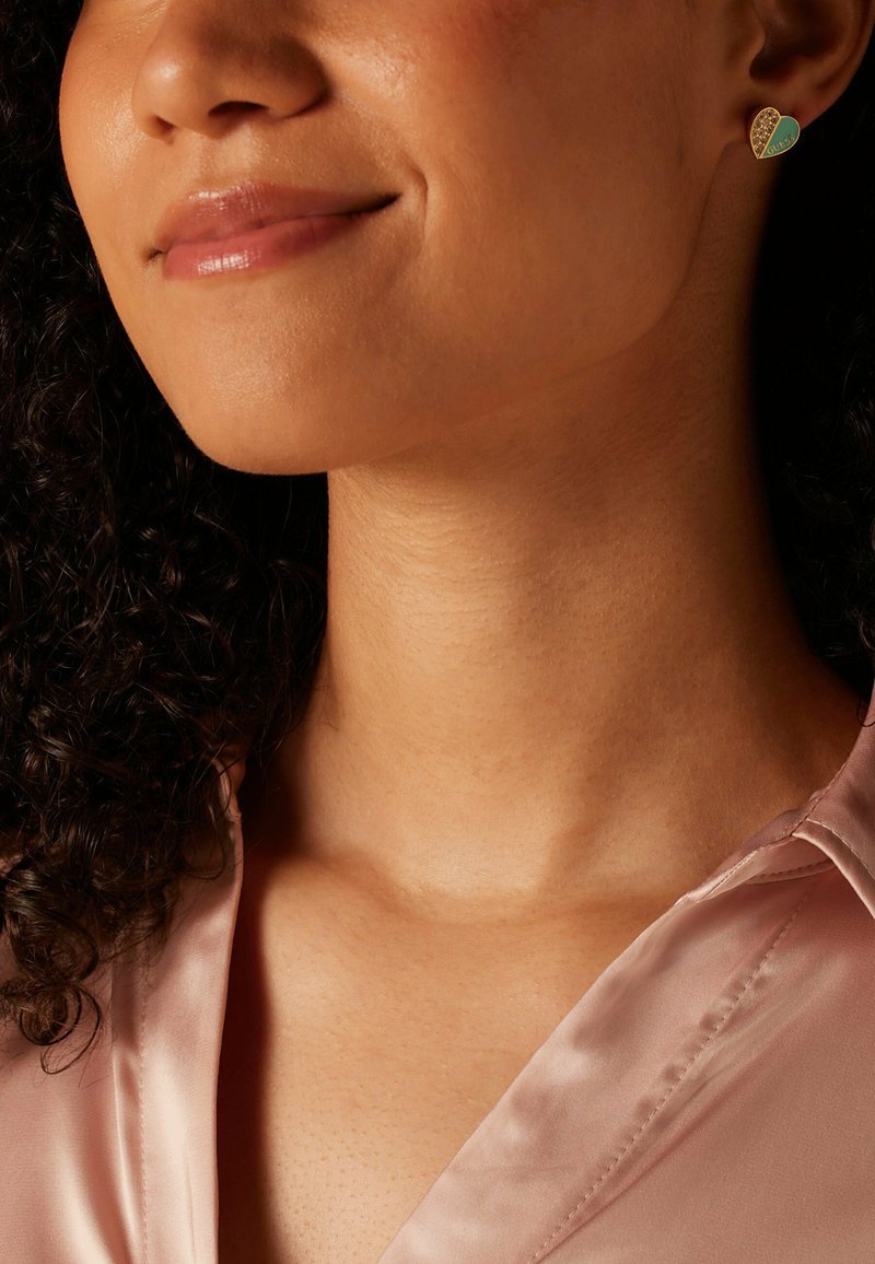 Boucles d'oreilles en forme de cœur en or avec un incrustation d'émail turquoise et un motif texturé. Le modèle porte un blouse rose clair ; une texture de peau subtile est visible.