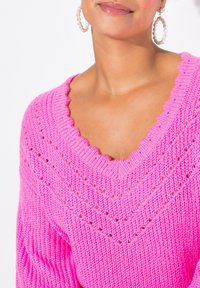 Pullover lavorato a maglia di un brillante colore rosa, con scollatura a V, caratterizzato da bordo scallop e motivi a traliccio. Texture a coste e vestibilità ampia. Il modello indossa orecchini a cerchio.