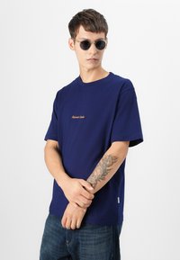 Camiseta azul marino de manga corta y cuello redondo, con un pequeño logo dorado en el frontal, combinada con jeans oscuros y gafas de sol.