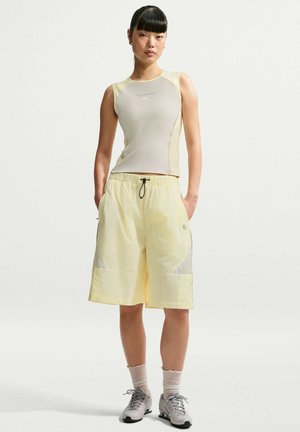 Vrouw staat met handen in de zakken, draagt een mouwloze beige top, losse gele shorts, lichte sokken en grijze sneakers op een witte achtergrond.