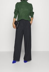 Pull côtelé vert à col roulé, pantalon noir à jambes larges, pochette noire et chaussures à talons hauts bleues. Matériaux texturés avec un design structuré.