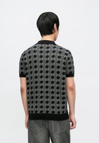 Čierna pletená polokošeľa so strieborným geometrickým vzorom, krátkymi rukávmi a rebrovaným golierom. Textília má hladkú textúru.