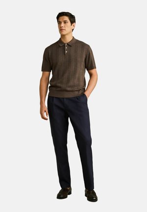 Man wearing a brown short-sleeve knit polo shirt, navy tailored pants, and dark brown dress shoes, standing with one hand in pocket.