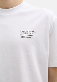 Balta medvilninė marškinėliai su spausdintu juodu dizainu, kuriame užrašyta "DUO TŪKSTANČIAI SEPTYNI - 2007" ir "DENIM TOM TAILOR" moderniu šriftu.