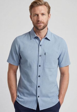 Mann mit Bart, der ein hellblaues Kurzarmhemd mit Brusttasche trägt, steht mit den Händen in den Taschen vor einem einfarbigen Hintergrund.