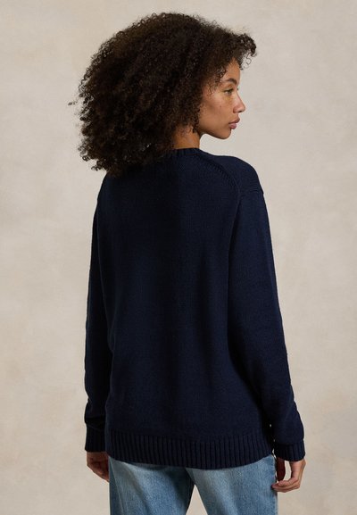 Woman with curly hair wearing a navy blue knit sweater and light blue jeans, facing sideways against a plain beige background.
