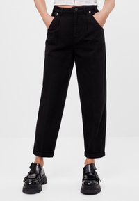 High-waisted black trousers with a loose fit, front pleats, side pockets, and rolled cuffs, paired with black platform shoes.