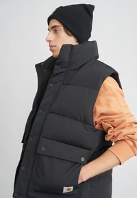 Svart puffväst med hög krage, tryckknappar och en framficka. Kombineras med en orange sweatshirt och en svart mössa.