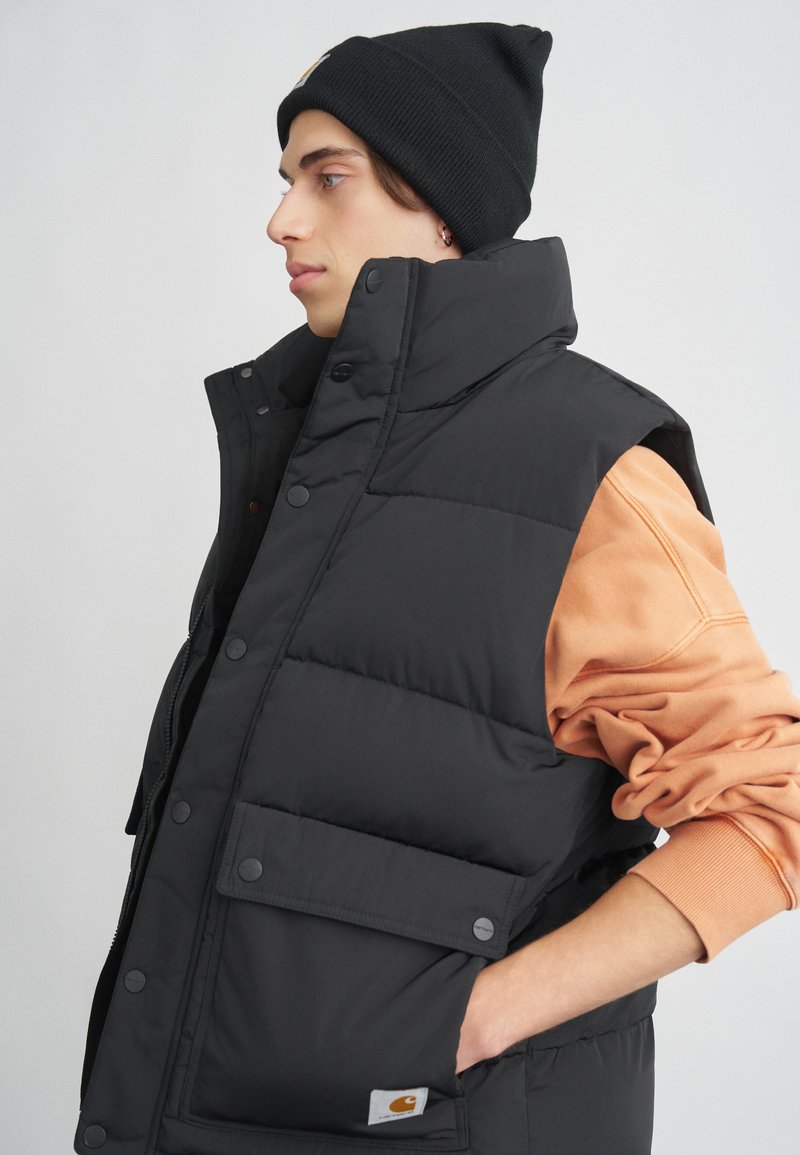 Svart puffväst med hög krage, tryckknappar och en framficka. Kombineras med en orange sweatshirt och en svart mössa.