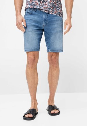 Mann trägt mittelblaue Jeansshorts, schwarze Slide-Sandalen und ein Hemd mit floralem Muster, steht auf weißem Hintergrund.