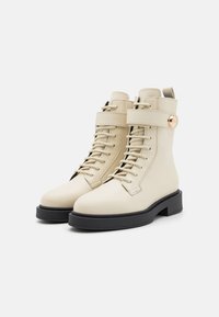 Beige skinnankler støvler med rund tå, snøring foran og sidebelte med gullakcent. Svart gummisåle og teksturert finish.
