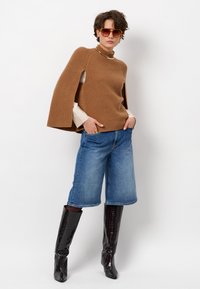 Bruine geribde trui met capesleeves, licht shirt eronder, blauwe denim culottes en zwarte glanzende kniehoge laarzen. Bruine zonnebril.