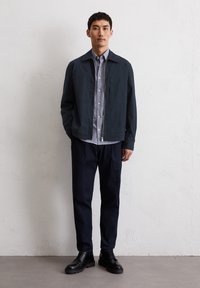 Navy lightweight jacket with a collared design, layered over a striped shirt, paired with dark trousers and black lace-up shoes.