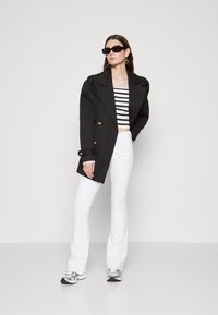 Svart doblet kneppet frakk, hvit stripete kort topp, hvite vide jeans og sølvfargede sneakers. Tilbehør inkluderer store svarte solbriller.