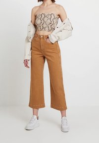 Haut court avec un motif serpent, associé à un jean ample taille haute marron et des baskets blanches. Toile de fond neutre.