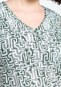 Tissu blanc avec un motif géométrique vert, doté d'un col en V et de deux boutons verts au niveau du col. Matériau texturé et léger.