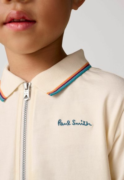 Krémová polo tričko s predným zipsom, rebrovaným golierom s modrými, ružovými a žltými pruhmi a vyšívaným nápisom "Paul Smith" v modrej farbe.