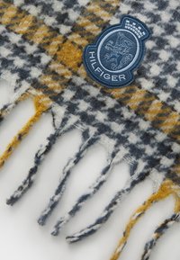 Wool scarf featuring a houndstooth pattern in navy, white, and mustard with fringed edges. Includes a blue embroidered logo patch.