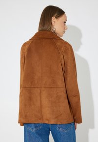 Braune Wildlederjacke mit Kragen, lässiger Schnitt und sichtbaren Nähten. Getragen über blauer Jeans, die eine glatte Textur aufweist.